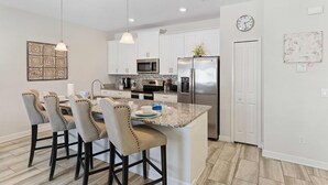 A modern kitchen with white cabinets, a stainless steel refrigerator, a microwave, and a granite island with bar stools.