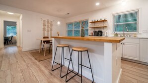 A kitchen with a wooden bar counter, two stools, a window with blinds, and a sink.