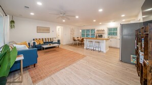A spacious living area with a sofa, coffee table, and a dining table with chairs. There is a kitchen area with a refrigerator, sink, and bar stools.
