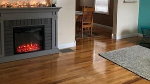 A living room with a fireplace, wooden flooring, a TV mounted on the wall, and a dining area in the background.