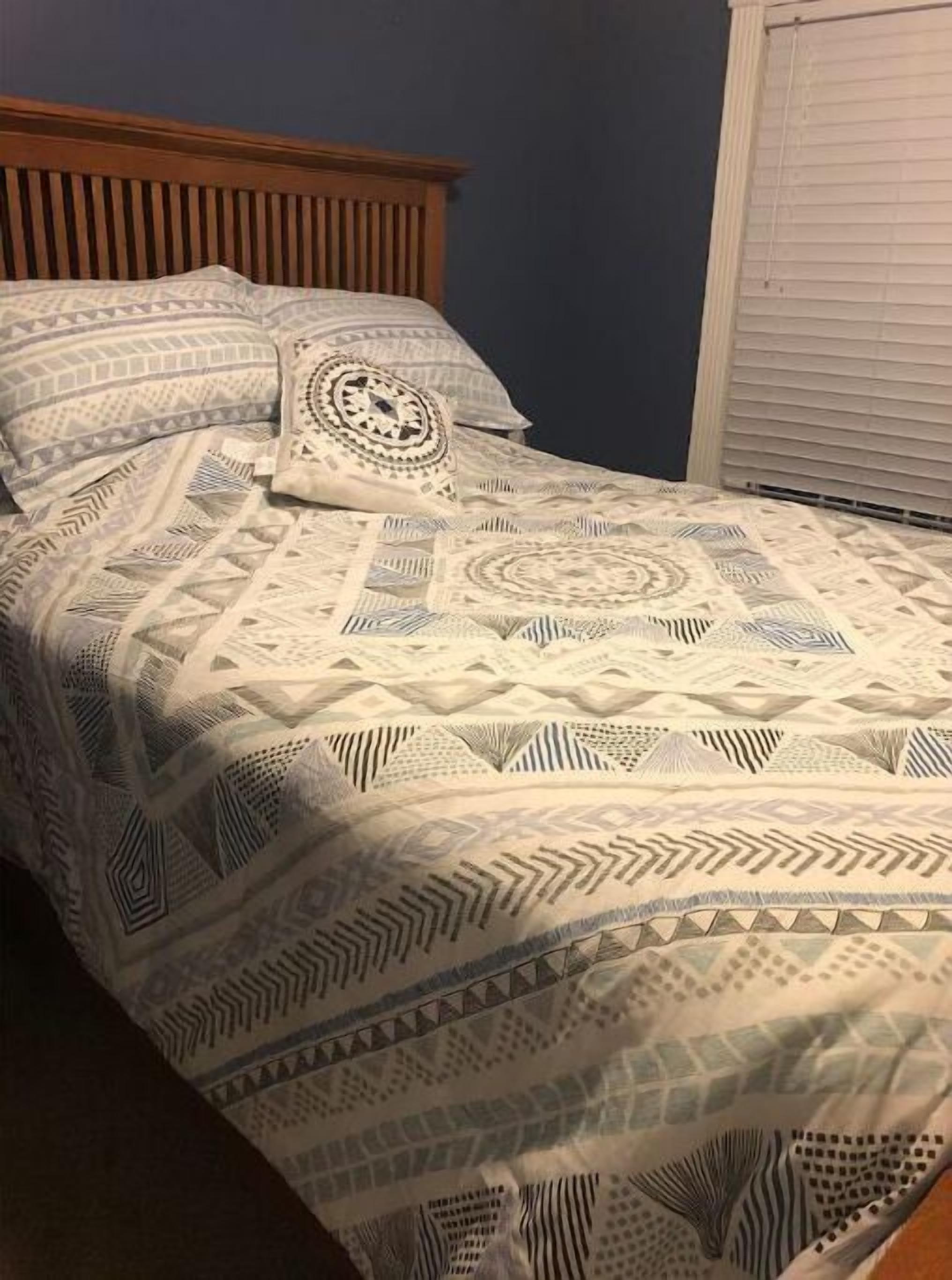 Una cama con colcha estampada y almohadas, cabecero de madera y ventana con persianas.
