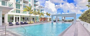 A modern hotel pool area with clear blue water, sun loungers, and a view of the city skyline. - Poolside District Condo - We Host (Miami)