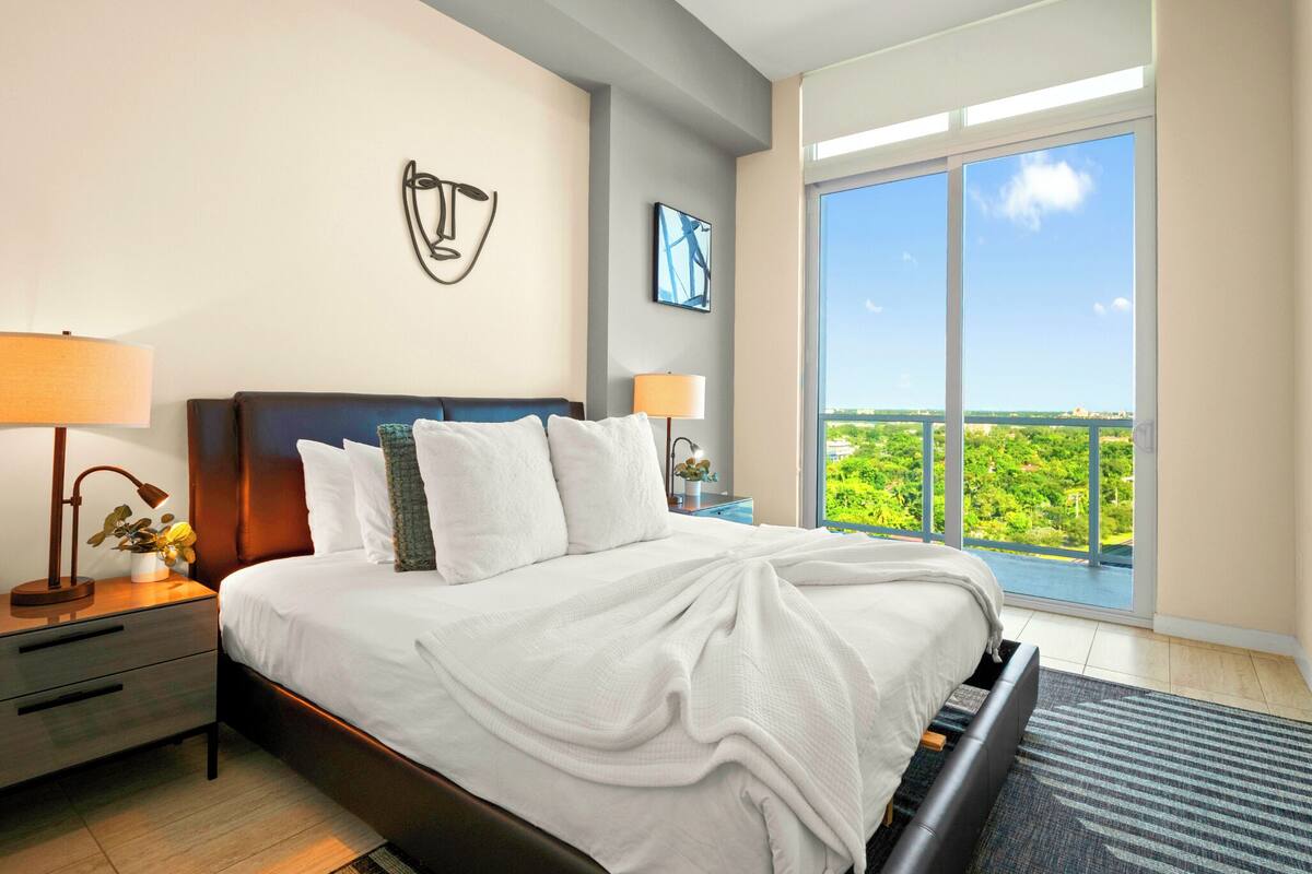 A modern hotel room with a large bed, a nightstand with a lamp, a wall art piece, and a large window offering a city view.