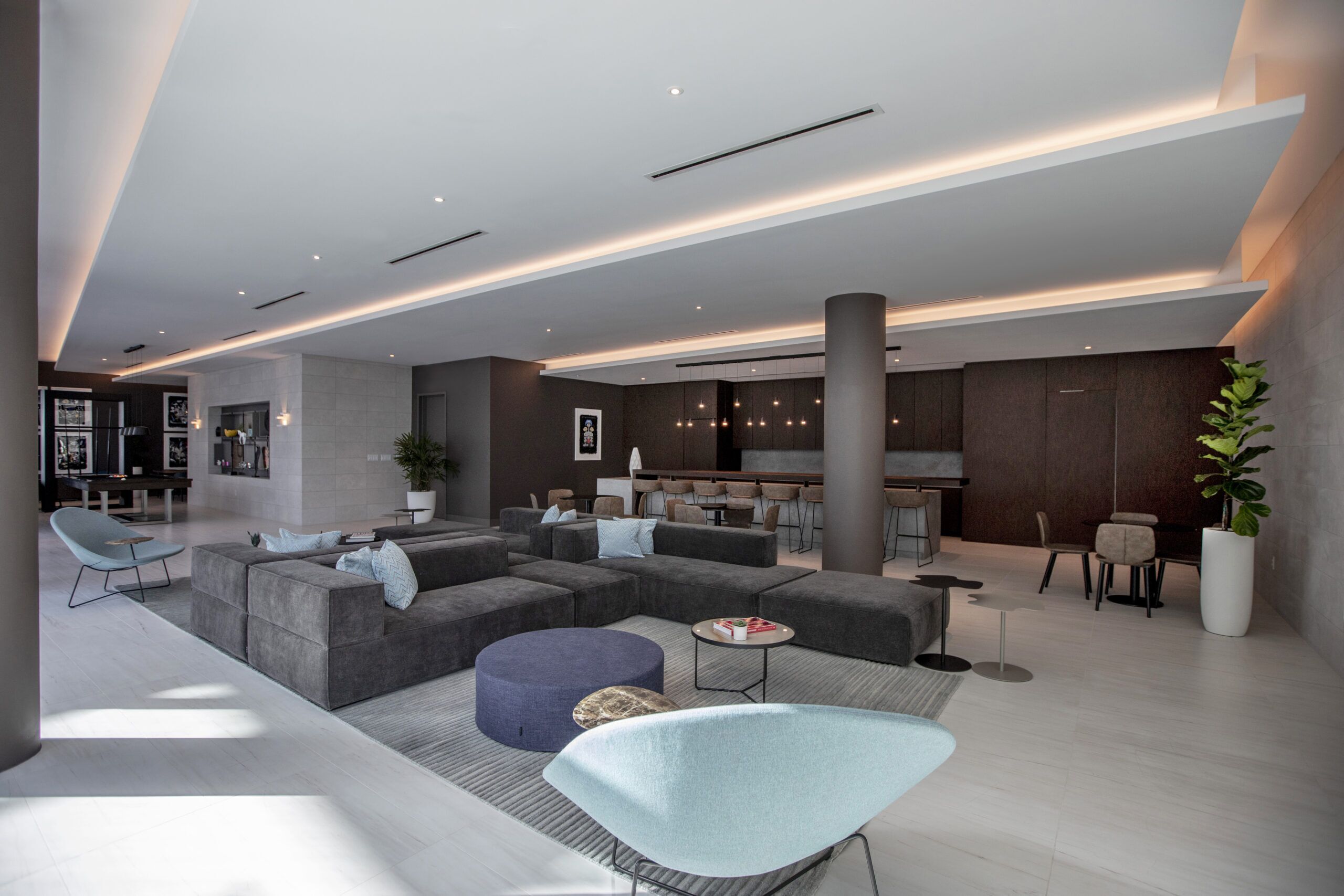 A modern living area with a sectional sofa, ottoman, and a unique chair. There is a kitchen area in the background with bar stools and a dining table.