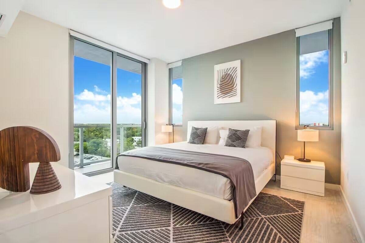 A modern bedroom with a large bed, a nightstand, a lamp, and a view of the sky through a sliding door.