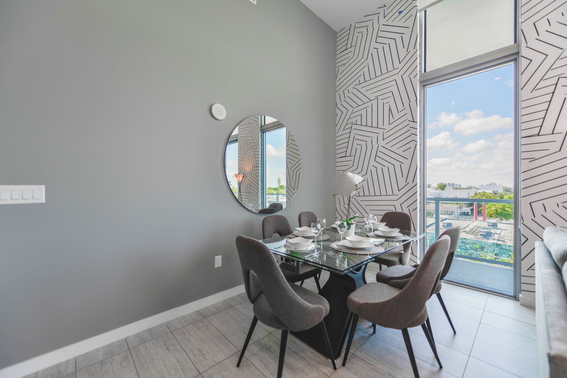 Un espace repas avec une table en verre, des chaises grises et un motif géométrique sur le mur.