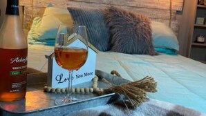 Un lit bien fait, recouvert d’une couverture à carreaux, une bouteille de vin blanc Arbol, un verre de vin et une carte sur laquelle est écrit : « Je t’aime plus que tout ».