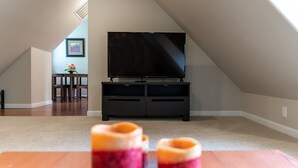 Une chambre mansardée avec un téléviseur à écran plat posé sur un meuble, une table à manger avec deux bougies et une petite table en bois sur laquelle est posé un vase de fleurs.