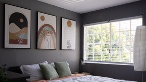 Une chambre moderne avec un grand lit, une fenêtre avec des stores et trois tableaux abstraits accrochés au mur.