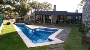 A modern house with a rectangular swimming pool, a patio with outdoor furniture, and a stone wall.