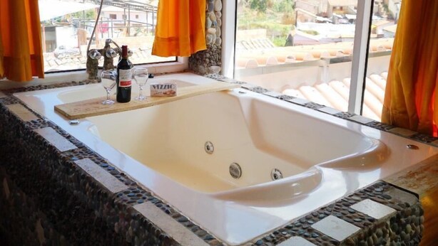 A bathroom with a large bathtub, stone mosaic tiles, and a view of a town through a window with orange curtains. - Cities Of the World - Cusco City Views (Cusco)