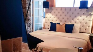 A bedroom with a tufted headboard, a large bed with blue and beige bedding, a bedside table with a mirror and a bottle of shampoo, and a view of the outside through a window with curtains.