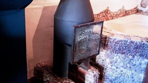 A stone fireplace with a metal chimney, logs, and a stone wall.