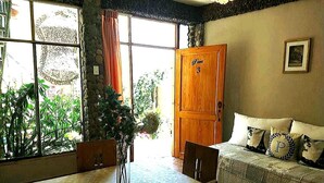 Una habitación con una mesa de comedor, sillas, un sofá con cojines decorativos, una puerta de madera y una ventana con plantas afuera.