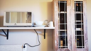 Una cocina con un microondas blanco, un cuenco blanco y una taza blanca sobre una estantería. Hay un mueble de madera con puertas de cristal que contiene botellas.