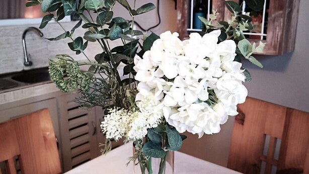 Una mesa con un jarrón de flores blancas y follaje verde.