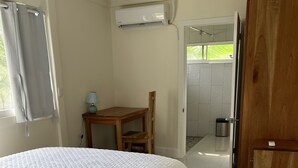 A hotel room with a large bed, a wooden table with a chair, an air conditioning unit, and a view of the outside.
