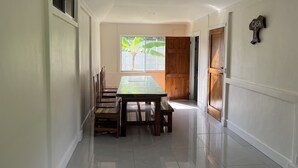 A hallway with a dining table and chairs, a window with a view of a banana tree, and a wooden door.