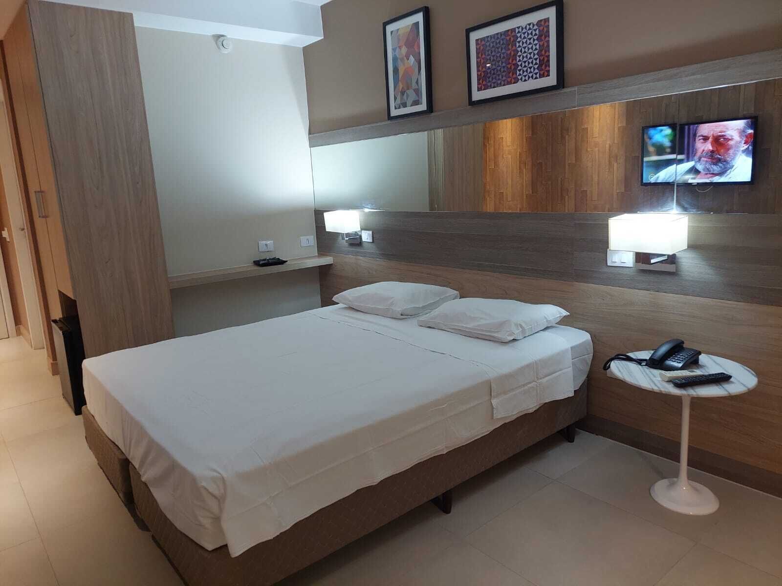 Una habitación de hotel con cama, mesitas de noche, televisión y teléfono sobre una mesita pequeña.