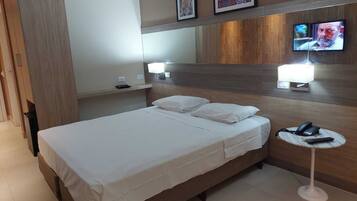 Una habitación de hotel con cama, mesitas de noche, televisión y teléfono sobre una mesita pequeña.