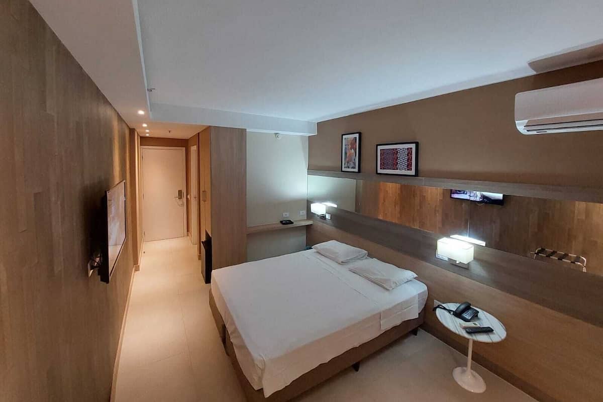 A modern hotel room with a wooden floor, a bed with white linens, a bedside table with a phone and remote, a wall-mounted TV, and a large mirror.