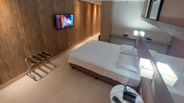 A modern hotel room with a wooden wall, a bed, a TV, a chair, and a small table with a phone and remote controls. - Mar Premier Flats (Queimados)
