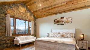 Une chambre avec un mur en pierre, un plafond en bois, un lit avec une literie à motifs, un canapé blanc et une table de chevet avec une lampe.
