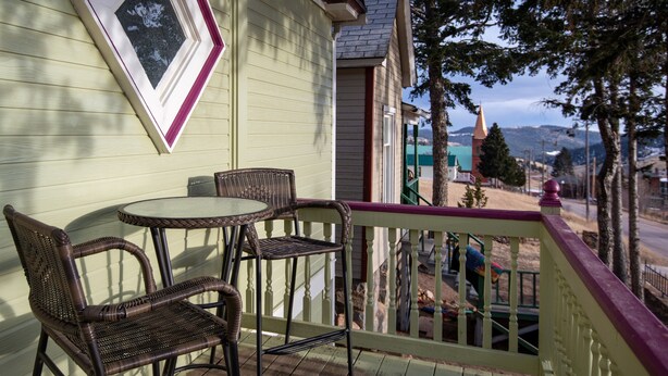 A balcony with wicker chairs and a table, overlooking a street with houses and a mountain in the background. - Central A/C1895 Victorian*Hot Tub*MTN Views (Cripple Creek)