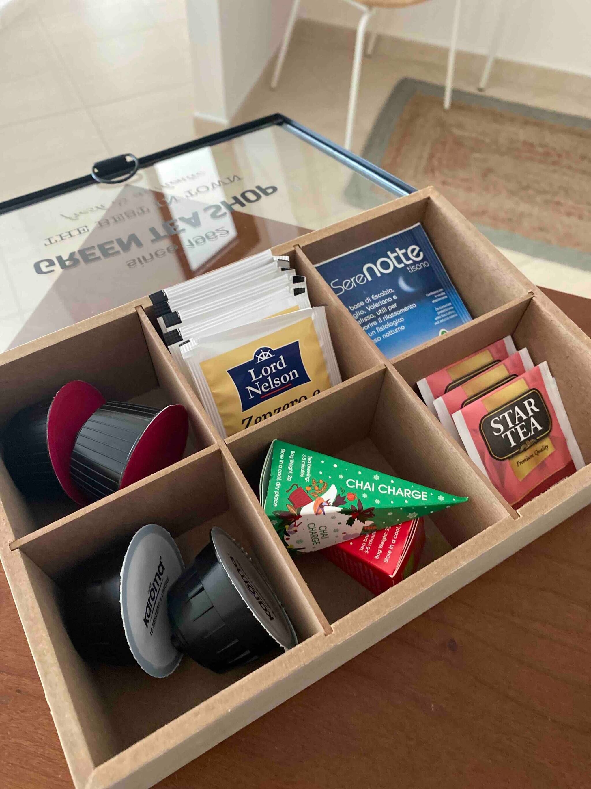 A wooden box with compartments containing various items including tea bags, a tea infuser, and a small decorative item.