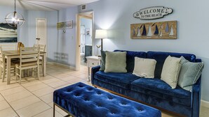 Une pièce avec un canapé en velours bleu, une table à manger en bois avec des chaises, un banc, une lampe et une pancarte indiquant «Bienvenue à la plage».