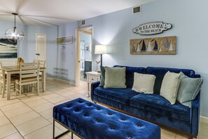Une pièce avec un canapé en velours bleu, une table à manger en bois avec des chaises, un banc, une lampe et une pancarte indiquant «Bienvenue à la plage».