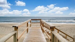 Une promenade en bois menant à une plage, avec un ciel dégagé et l’océan en arrière-plan.