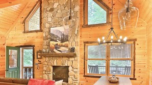 A cozy cabin interior with a stone fireplace, wooden walls, a dining area, and a kitchen with a gas stove. - Luxury ADK Retreat Near Whiteface, Lake Placid (Jay)