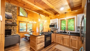 A rustic cabin interior with a stone fireplace, wooden ceiling beams, a kitchen with a large island, and a dining area. - Luxury ADK Retreat Near Whiteface, Lake Placid (Jay)