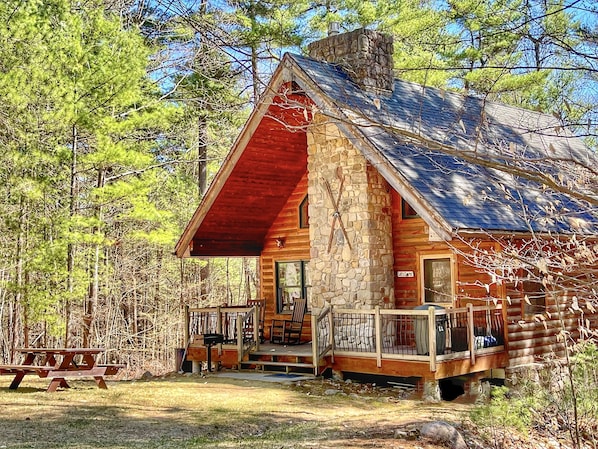 A rustic cabin with a stone chimney, wooden exterior, and a porch with a railing.