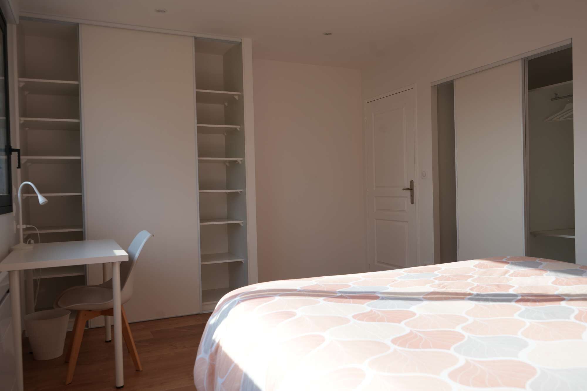 Een slaapkamer met een bed, een bureau en een stoel. Er is een garderobe met meerdere planken en een deur.