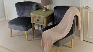 Dos sillones grises oscuros con patas de madera, una mesita auxiliar color beige con una lámpara y un plaid de punto beige cubriendo uno de los sillones.