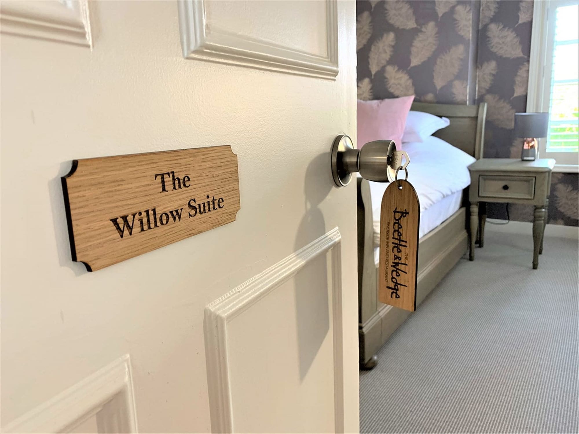 A hotel room with a bed, a bedside table, a lamp, and a sign on the door reading "The Willow Suite".