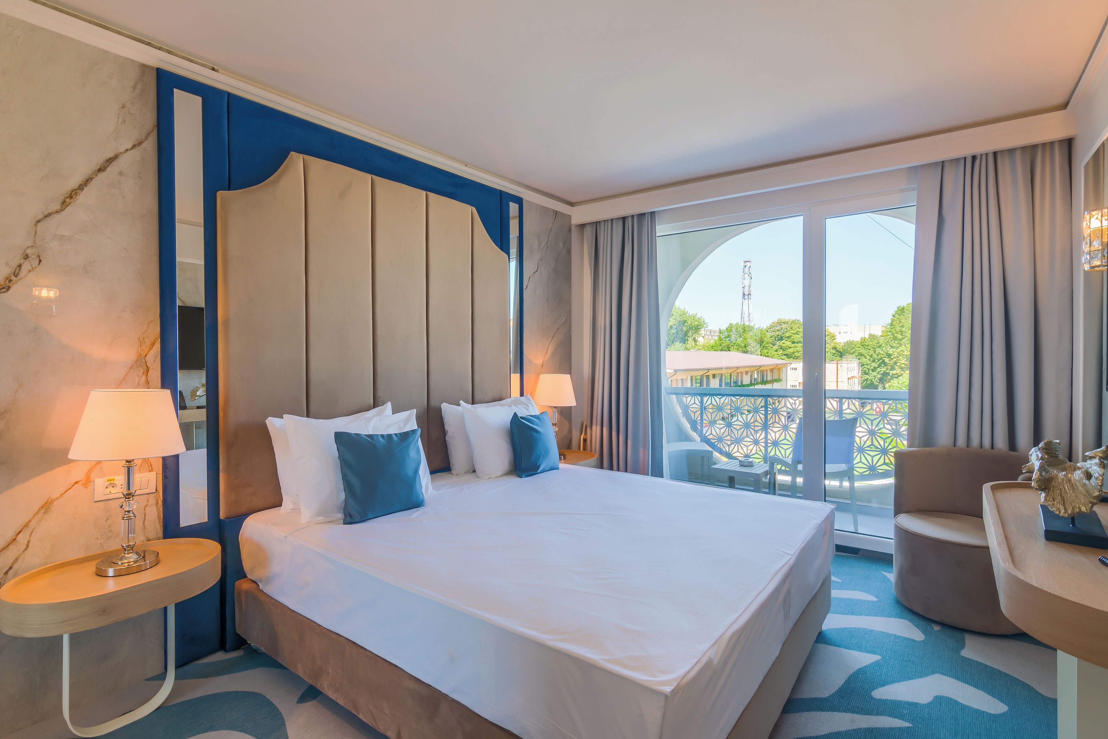 A hotel room with a large bed, two bedside lamps, a view of the outdoors, and a balcony with a chair.