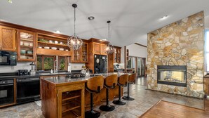 A modern kitchen with wooden cabinets, a large island with bar stools, a stone accent wall, and a built-in fireplace. - 14 Acres - Theater Room - 20 Miles to DT Asheville - Paved Drive the whole way! (Canton)