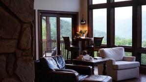 A cozy living room with a stone wall, a leather sofa, a white armchair, and a view of a lush landscape through large windows. - 14 Acres - Theater Room - 20 Miles to DT Asheville - Paved Drive the whole way! (Canton)