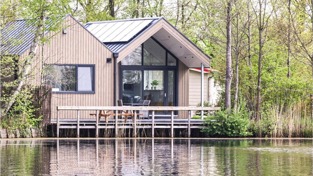 A modern cabin with a deck overlooking a calm lake, surrounded by trees.