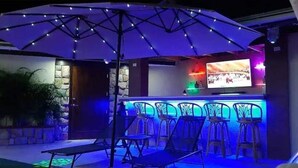 An outdoor bar area with a TV screen, multiple bar stools, and a table under a large umbrella. - Arupar Apartment (PARADERA)