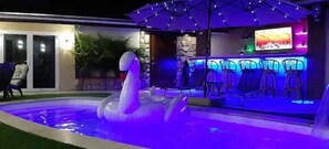 Une piscine avec un flotteur en forme de cygne, un coin bar avec des chaises et un écran de télévision.