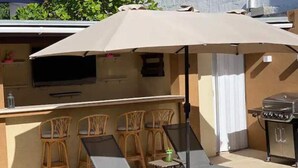 An outdoor patio with a barbecue grill, lounge chairs, and a table under a large umbrella. - Arupar Apartment (PARADERA)