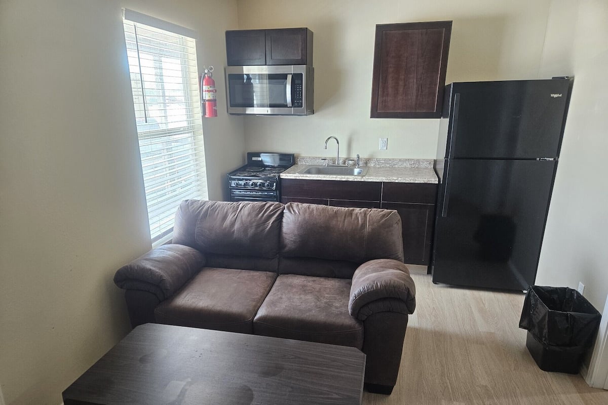 A living room with a brown sofa, a dark wooden coffee table, a kitchen with a stove, microwave, sink, and black refrigerator.