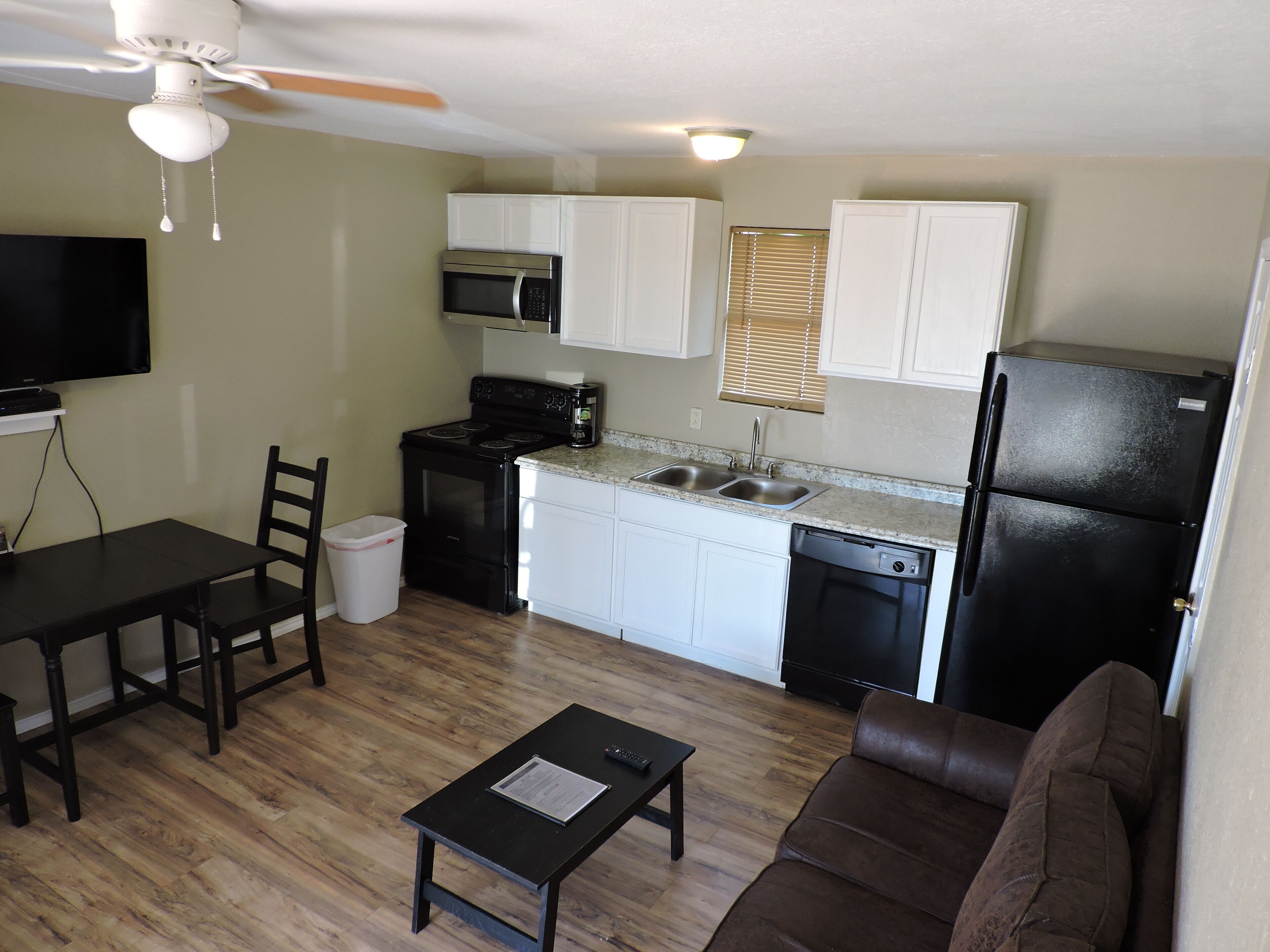 A compact kitchen with white cabinets, a black refrigerator, and a microwave. There's a dining table with chairs, a sofa, and a small table with a remote control.