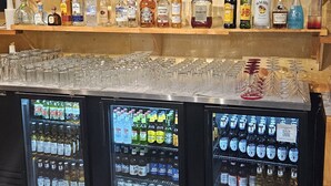 Zona de bar con estantes de botellas de licor y vasos, y dos vitrinas refrigeradas con cerveza y otras bebidas.