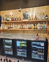 Zona de bar con estantes de botellas de licor y vasos, y dos vitrinas refrigeradas con cerveza y otras bebidas.