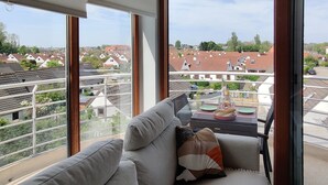 En balkon med sofa og spisebord, med udsigt over et boligområde med røde tegltage.